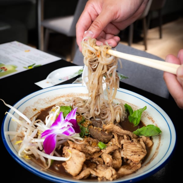 Hands using chopsticks to lift noodles from a bowl of Thai boat noodles with meat and vegetables.