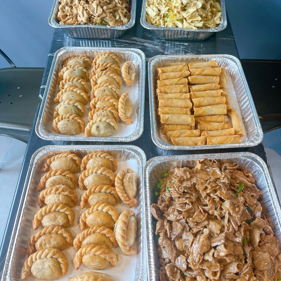 Four aluminum trays filled with various Asian dishes, including empanadas, spring rolls, and stir-fried meat.