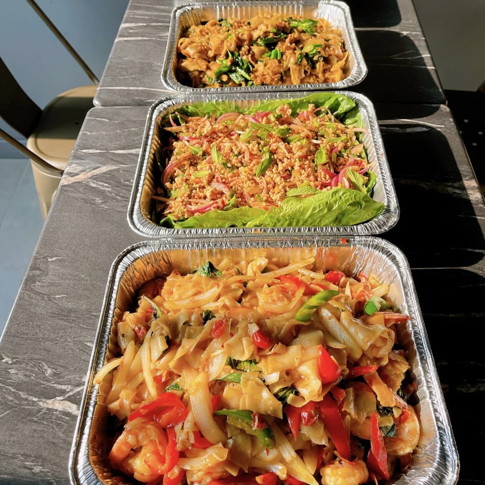 Three foil trays of Thai food, including noodles with shrimp and vegetables, and a minced meat salad.