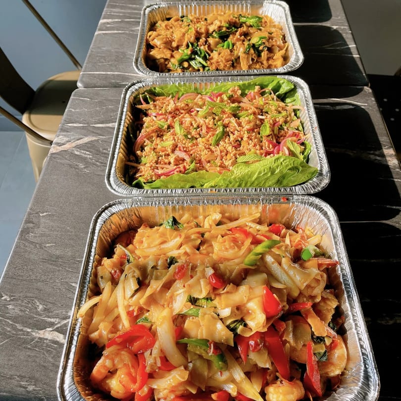 Three foil trays of Thai food, including noodles with shrimp and vegetables, and a minced meat salad.