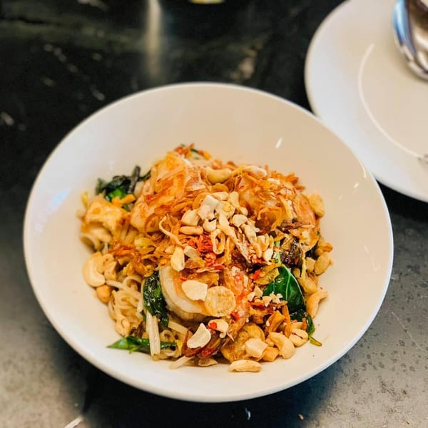 A white bowl filled with noodles, shrimp, cashews, fried onions, and green herbs on a dark table.