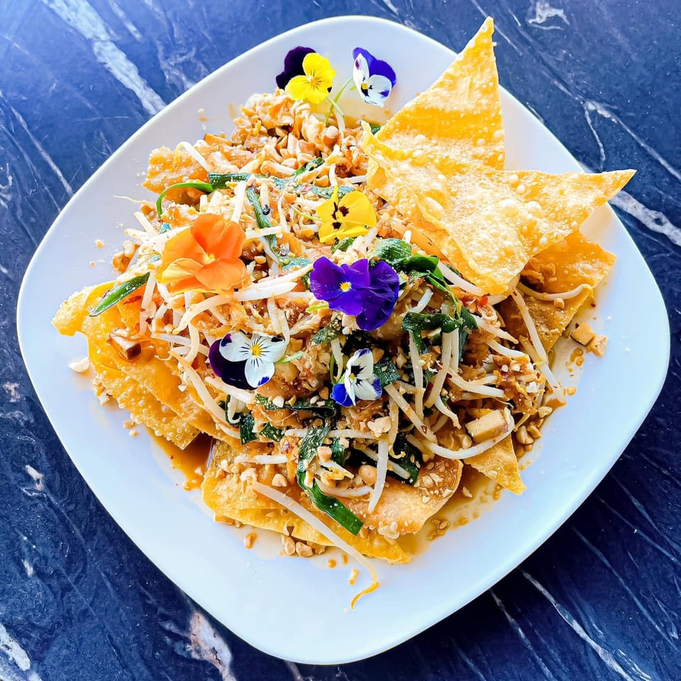 Pad Thai with fried wontons, bean sprouts, peanuts, and edible flowers on a white plate.