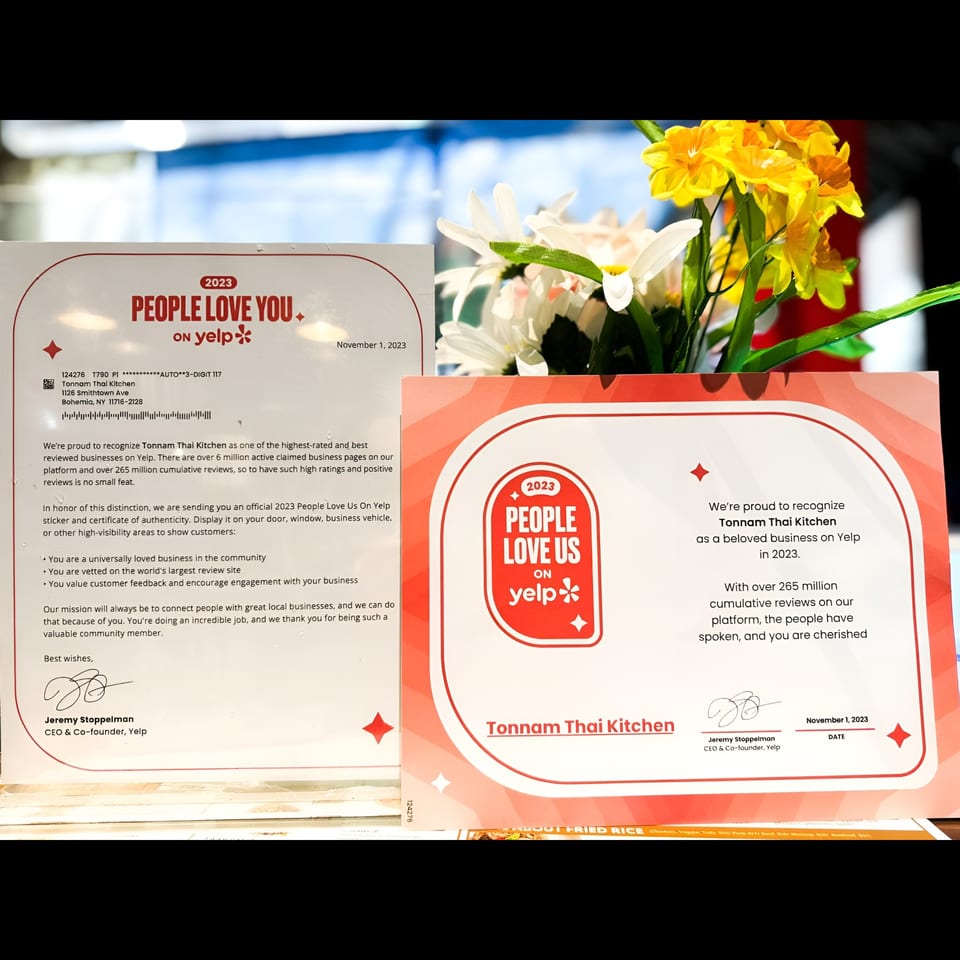 Two Yelp "People Love Us" awards for Tonnam Thai Kitchen sit on a table with flowers.