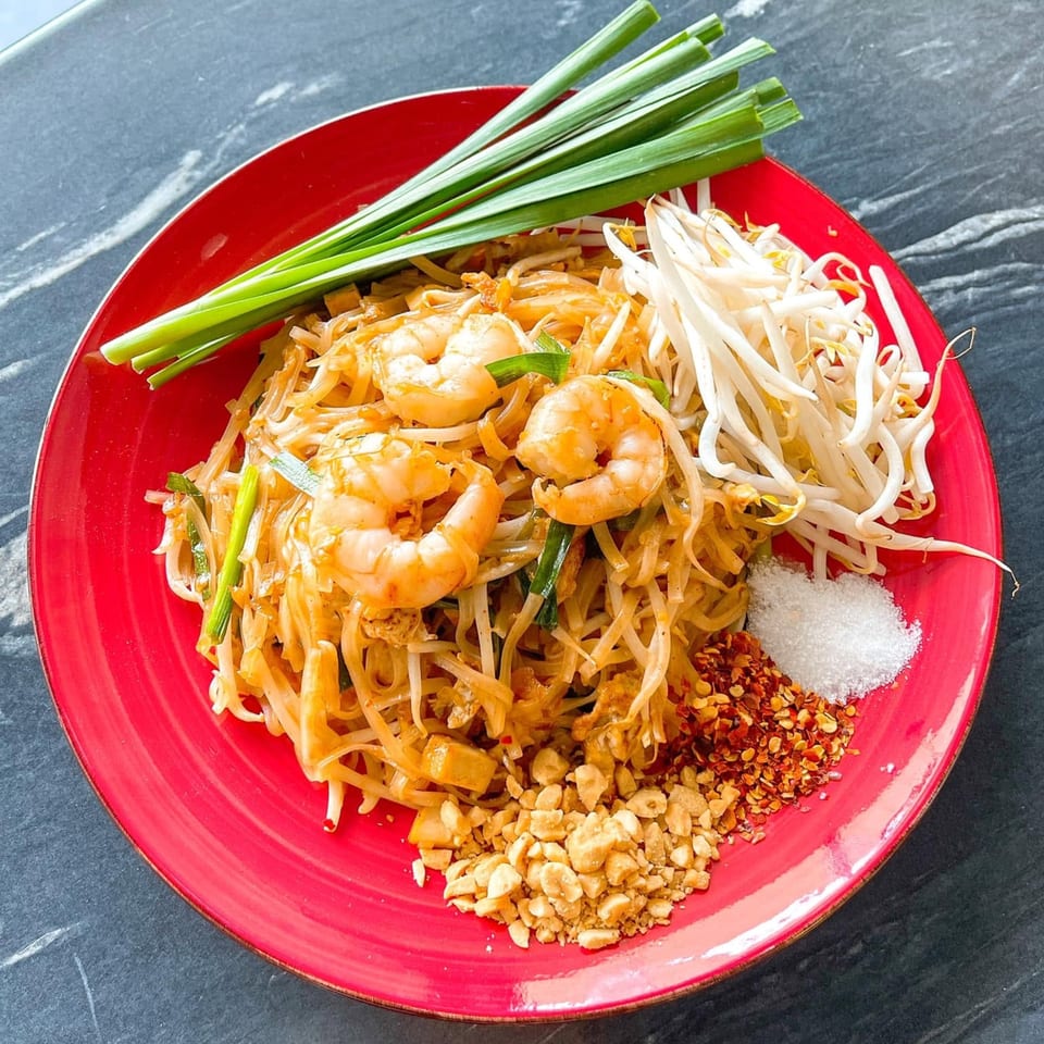 Shrimp Pad Thai with bean sprouts, chives, crushed peanuts, chili flakes, and sugar on a red plate.