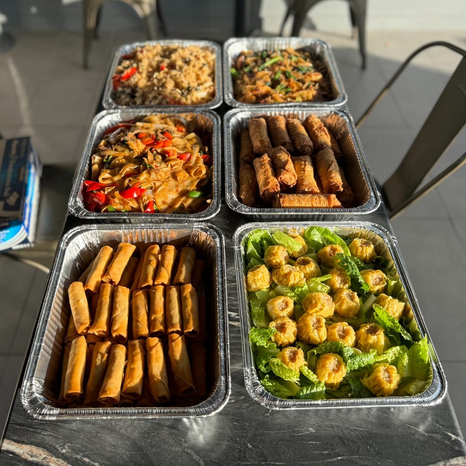 Six foil trays of Asian catering food, including fried rice, noodles, spring rolls, and dumplings.
