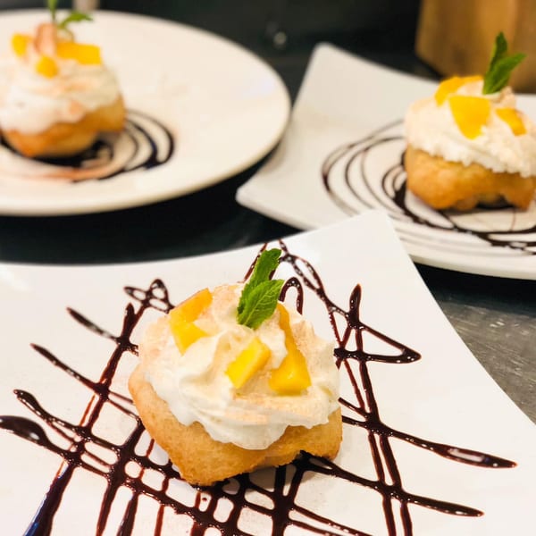 Three golden-fried desserts topped with whipped cream, mango, and mint, drizzled with chocolate sauce.