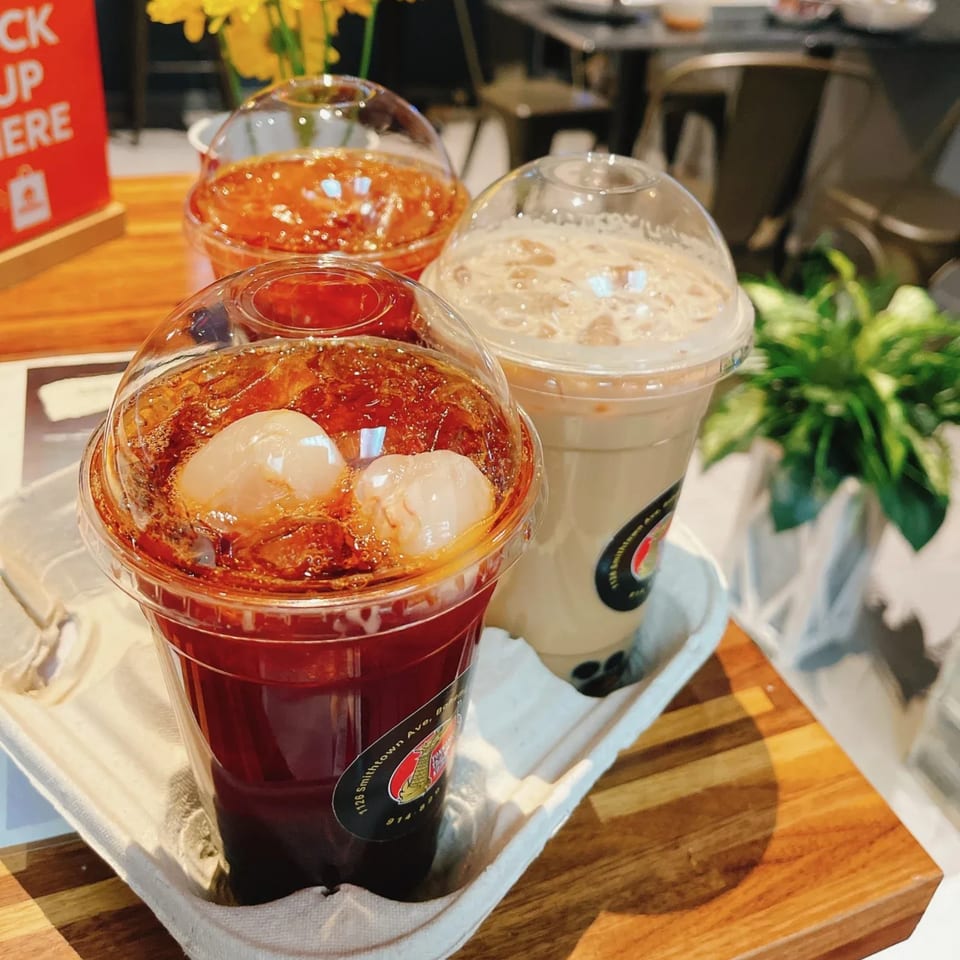 Three plastic cups of iced beverages, two red with lychee and one creamy, on a wooden table.