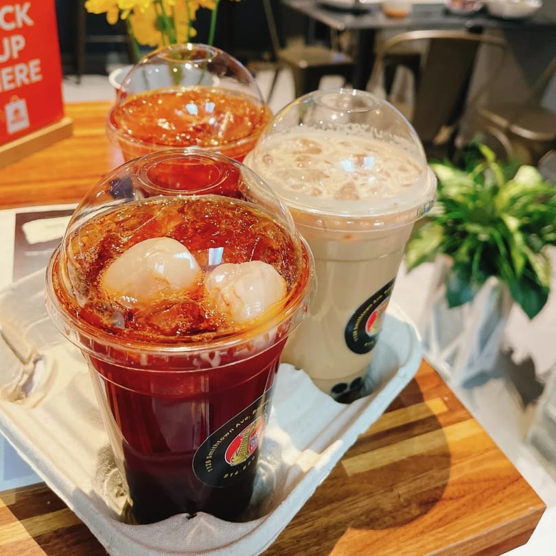 Three plastic cups of iced beverages, two red with lychee and one creamy, on a wooden table.