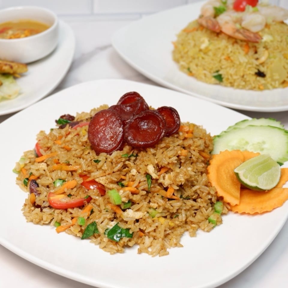 Fried rice with sliced sausage, vegetables, lime, and cucumber, with another plate of fried rice in the background.