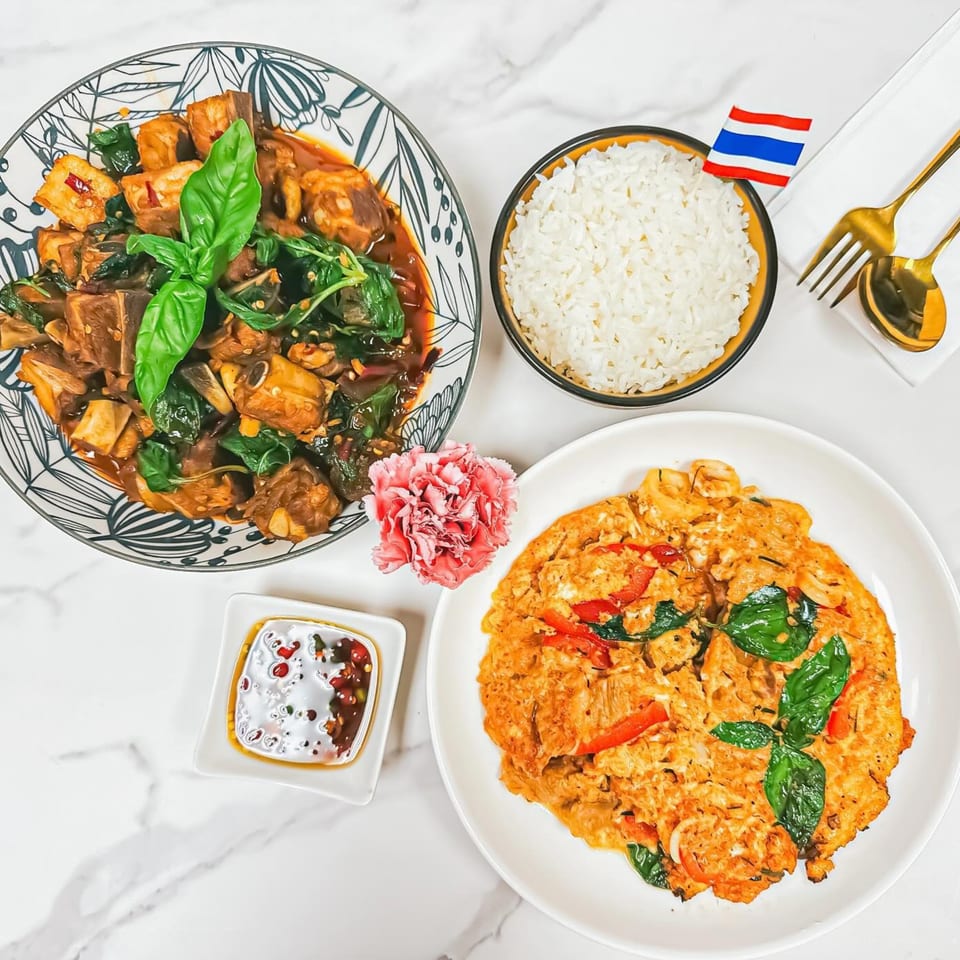 Thai basil pork ribs, omelet with basil, rice, chili sauce, and a Thai flag on a marble table.