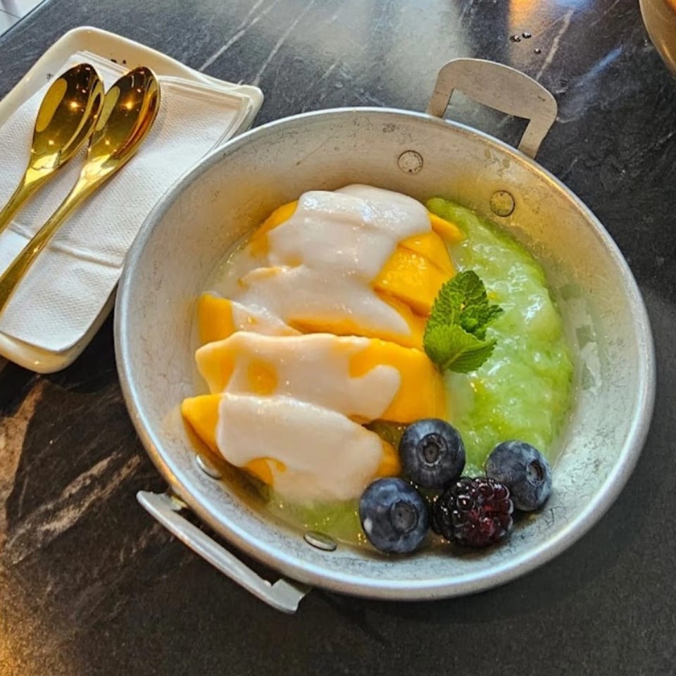 Mango sticky rice dessert with coconut cream, green rice, blueberries, and mint in a metal bowl.