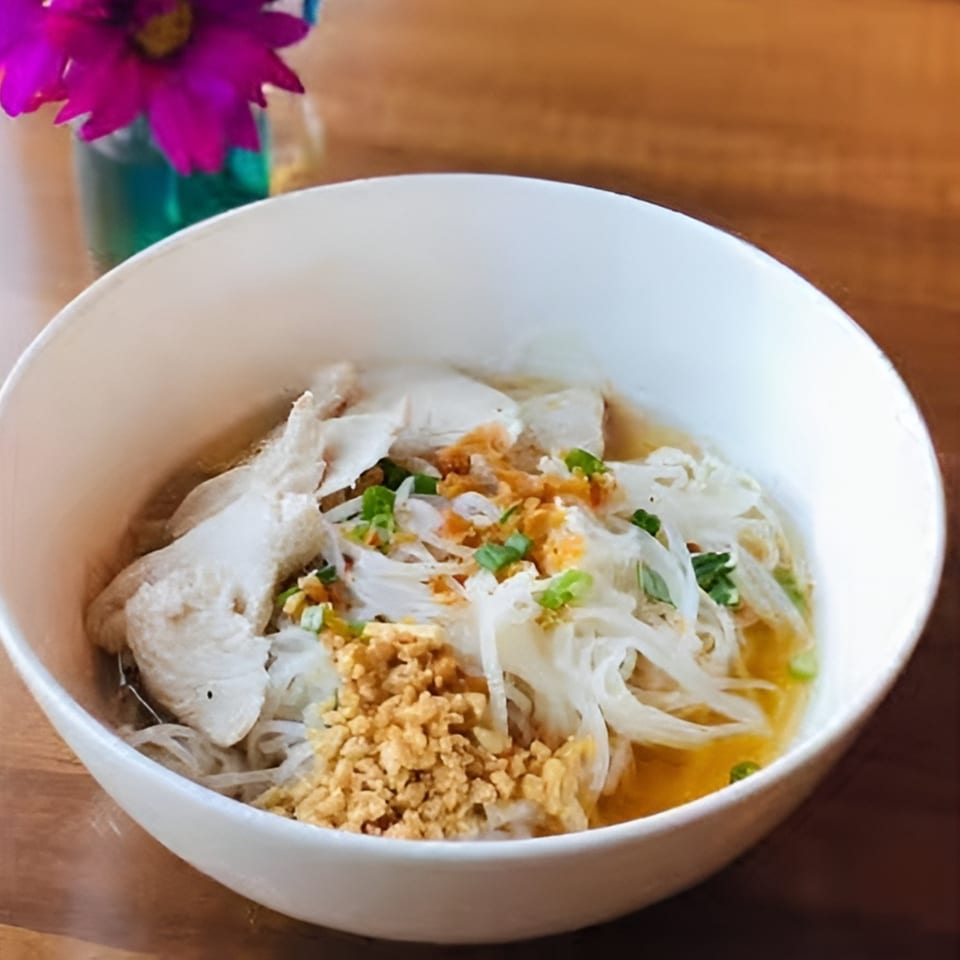 A white bowl of chicken noodle soup with green onions and crispy garlic on a wooden table.