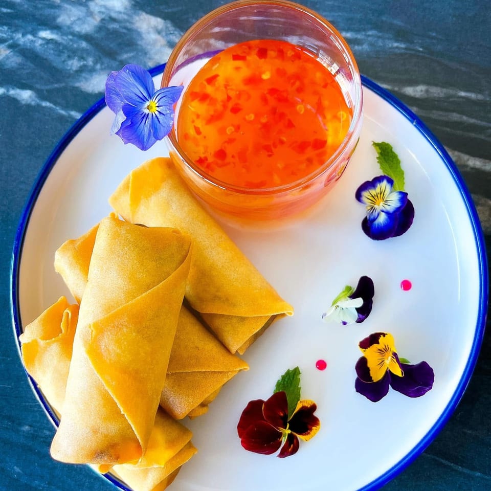 Crispy golden spring rolls with sweet chili dipping sauce, garnished with edible flowers on a white plate.